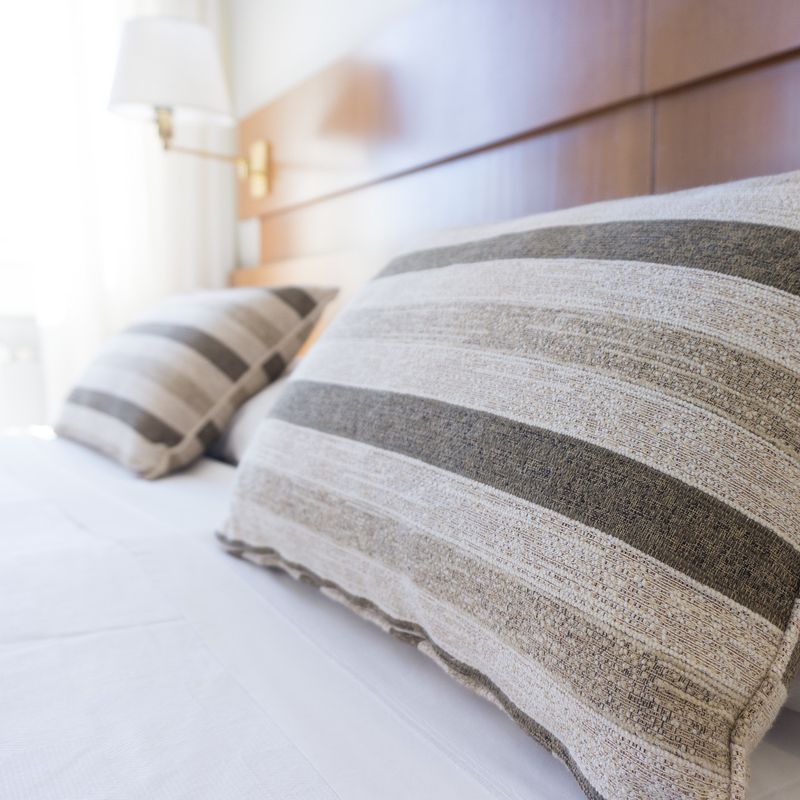 Close-up of a neatly made bed with two striped throw pillows in neutral tones, resting against a wooden headboard in a bright, sunlit room. Image for Contact Rochelle Cleaners.